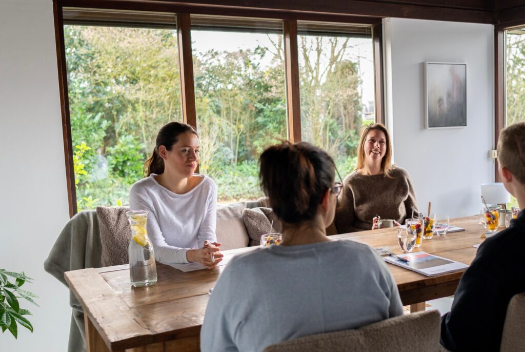 Een groep in gesprek aan de grote tafel bij Doyo, met de tuin achter de ramen