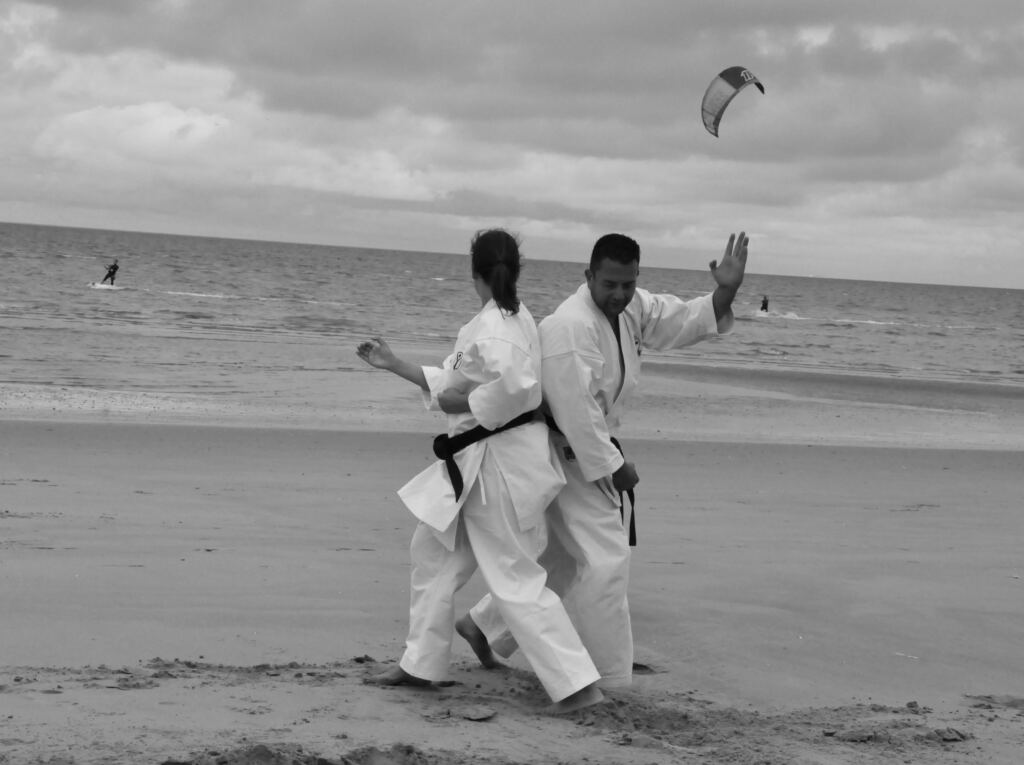 Chantal en Arno in karatepak met zwarte band, rug aan rug op het strand, met kitesurfers op zee