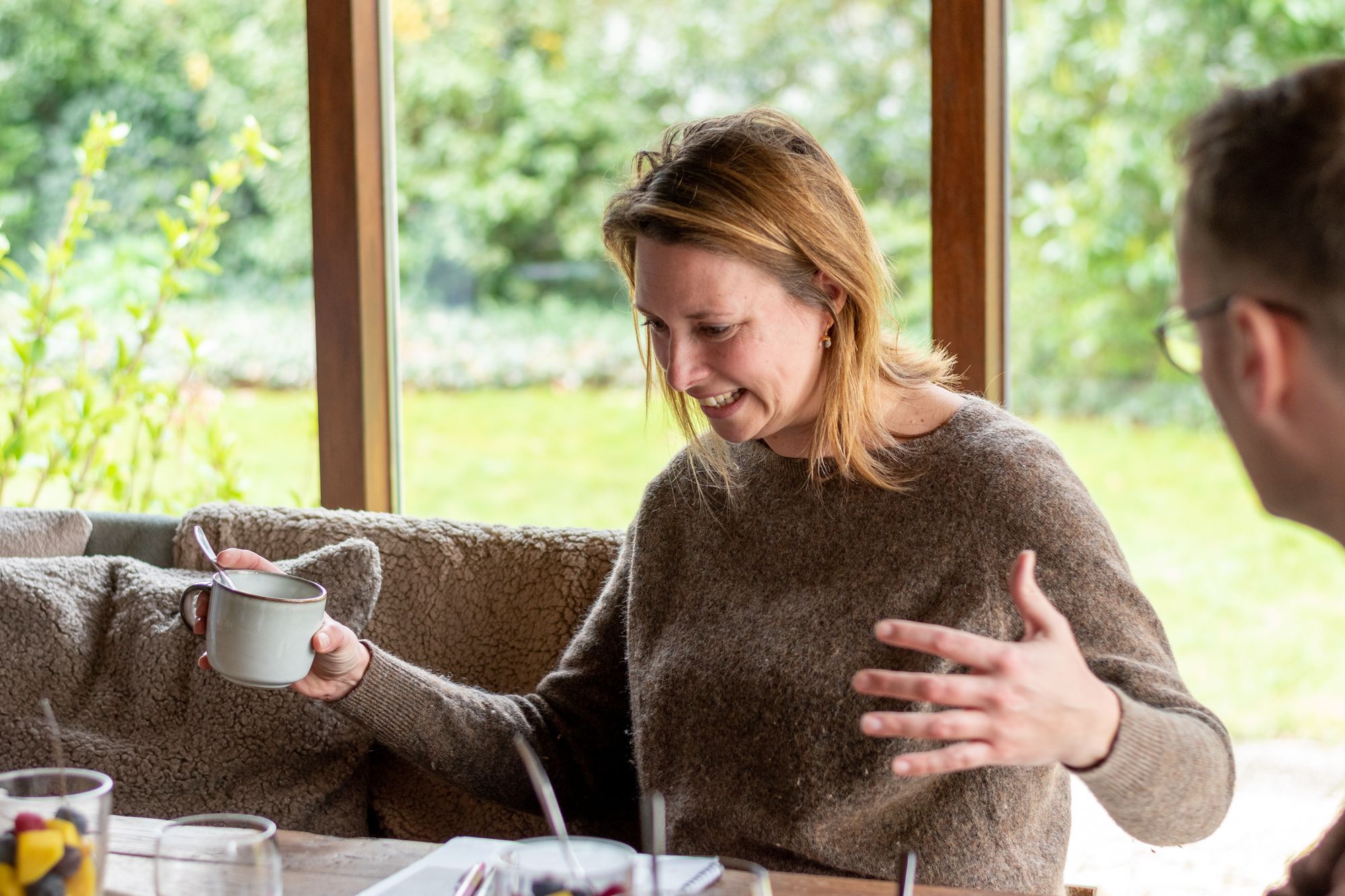 Deelnemer lacht en vertelt met een kop koffie in de hand aan de grote tafel bij Doyo, met de tuin achter het raam